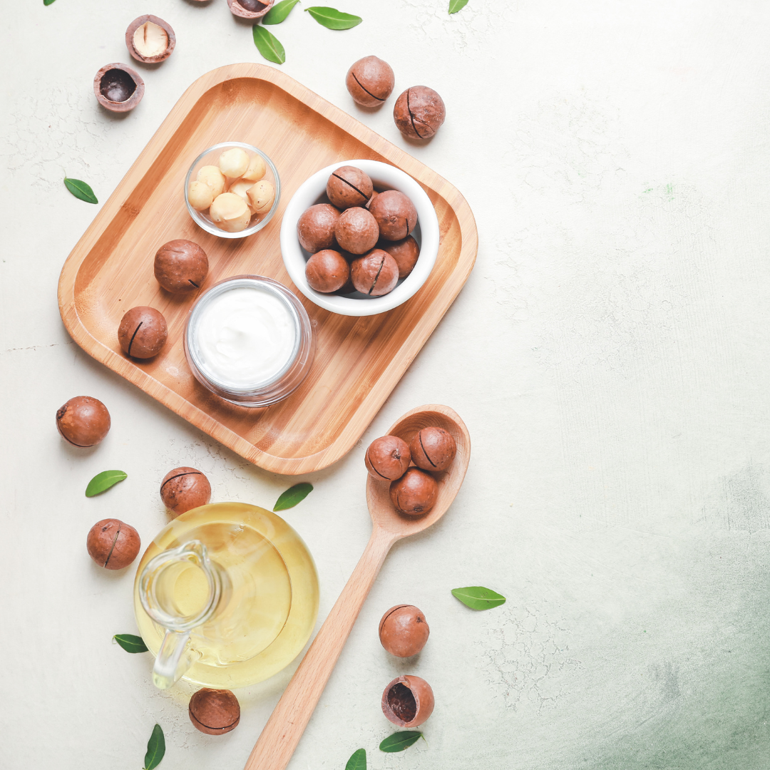 Macadamia nuts, oil, and a wooden spoon on a light surface with green leaves.